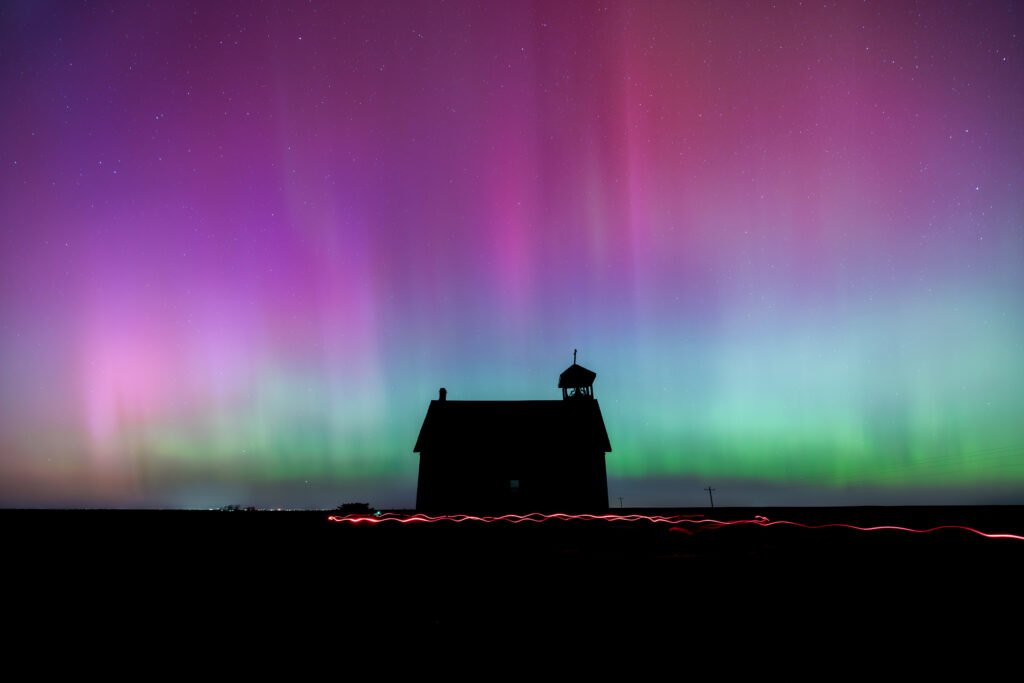 The northern lights shining in purple, pink, blue, and green above Abbott Church in the middle of the night. The dark field in front of the church has a red light streaked across the ground.