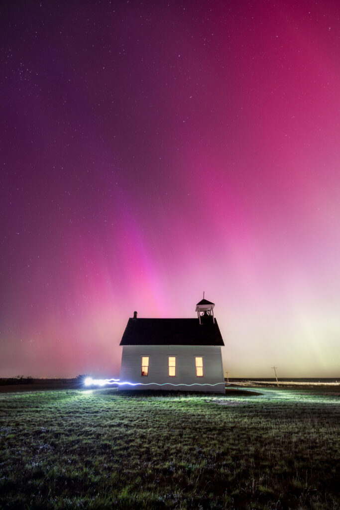 The northern lights shining in purple, pink, yellow, and green above Abbott Church in the middle of the night. The field surrounding the church is painted bright with a white light showing green and yellow blades of grass.