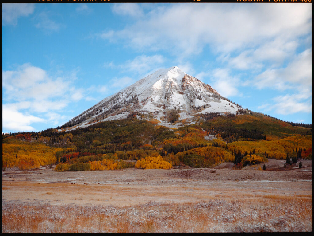 A snow-capped Gothic Mountain under a bright blue sky. There are groves of yellow, green, and red aspens all around it, and a snowy field in the foreground