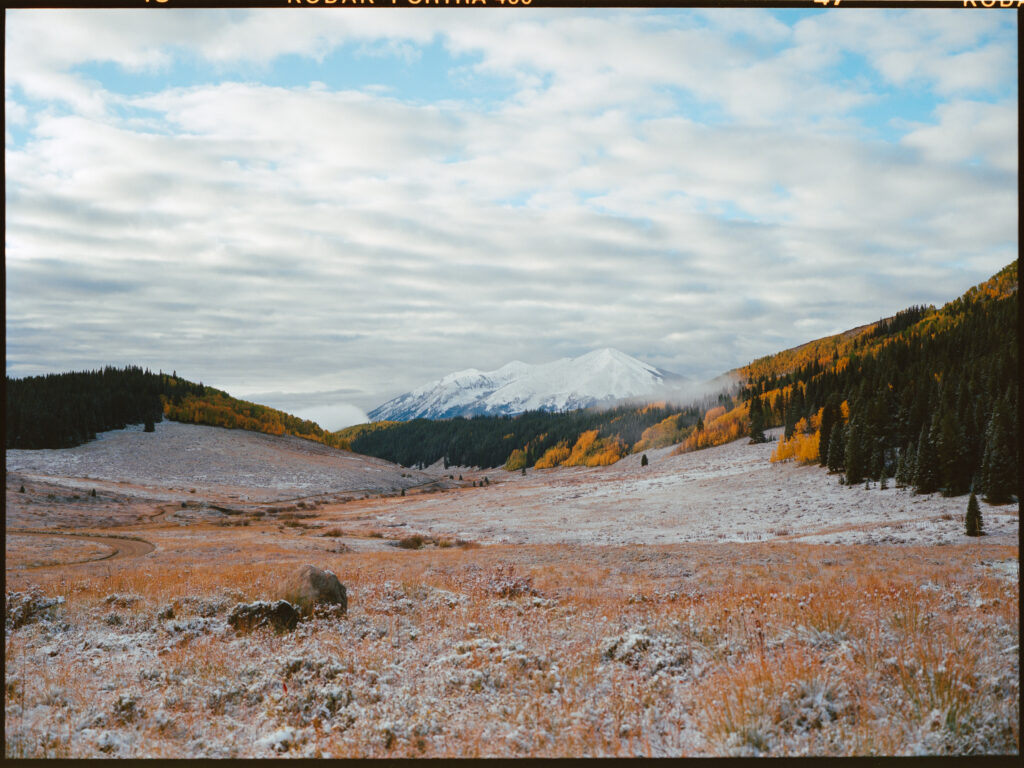 A snow-covered mountain above a valley of orange and yellow fall colors, pine trees, and covered in a layer of frost